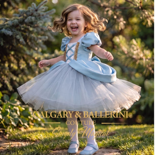 A young girl wearing a light blue and white tutu dress with gold trim is captured mid-spin outdoors in a grassy garden setting. The dress features puffed sleeves, a lace-up bodice, and a voluminous tulle skirt.