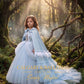 A young girl stands in a misty forest wearing a voluminous light blue gown with a matching cape and white feather trim. The image has a warm, sunlit magical quality, with a watermark reading 'CALGARY & BALLERINE COUTURE Simply Magical' at the bottom.