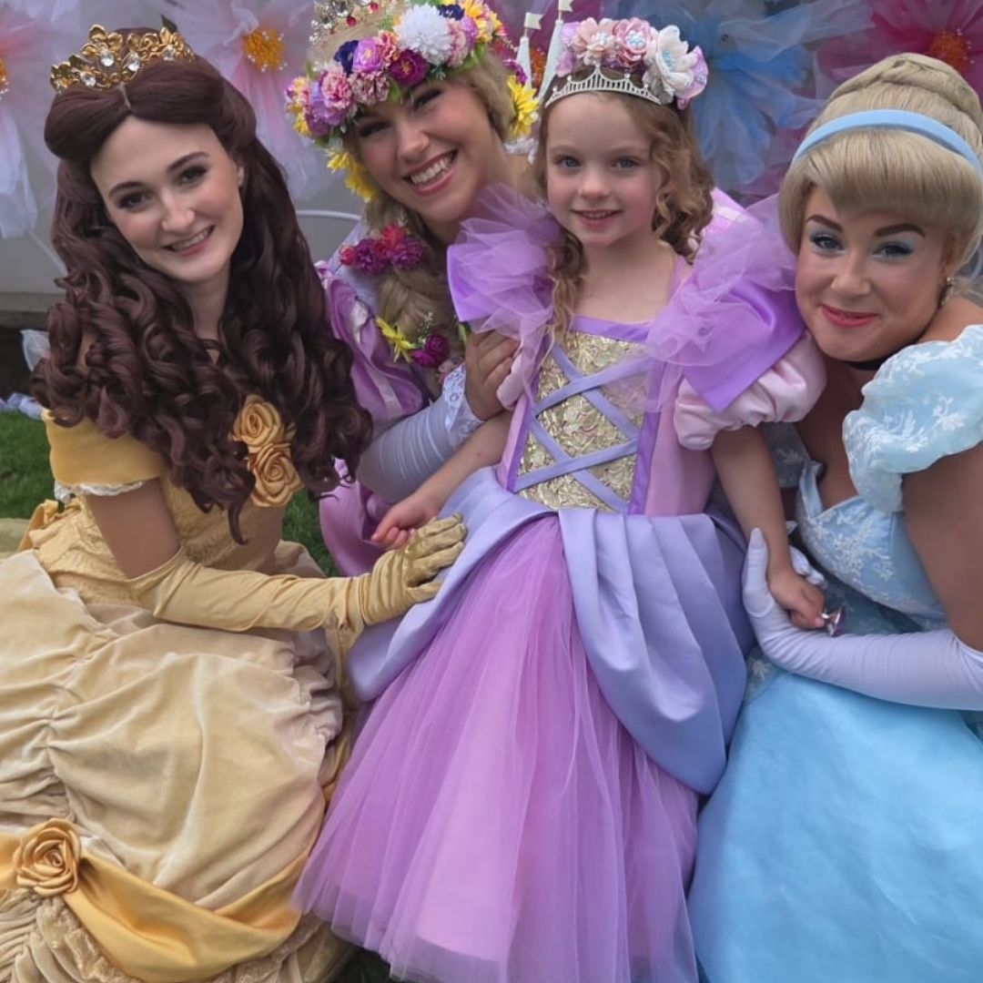 Four little girls in princess costumes posing together outdoors at a birthday party .