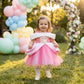 A young girl stands on grass outdoors wearing a pink and white princess-style dress with a voluminous tulle skirt and a white cape. Balloons, a flower arch, and warm sunlight are visible in the background.
